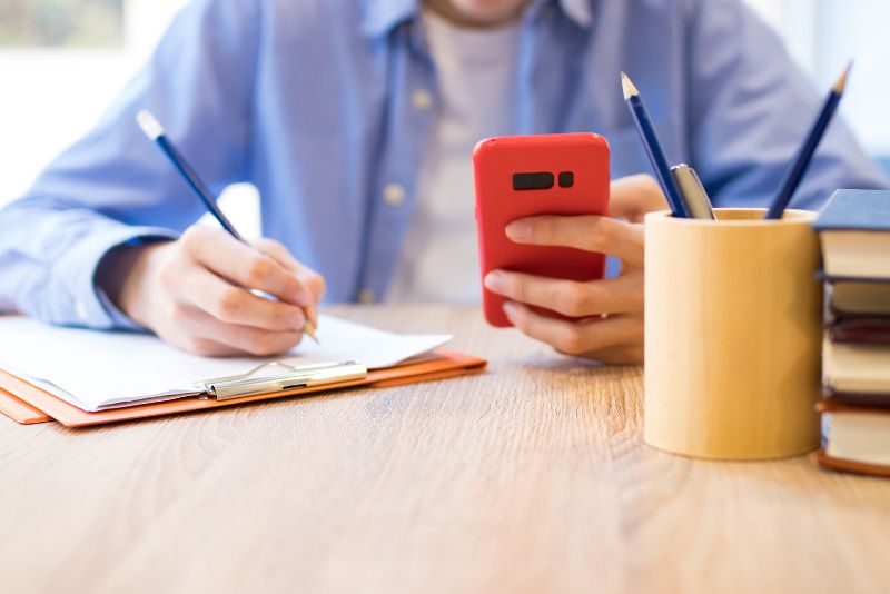 A man sitting at a desk taking notes with his right hand while holding his smartphone with the left