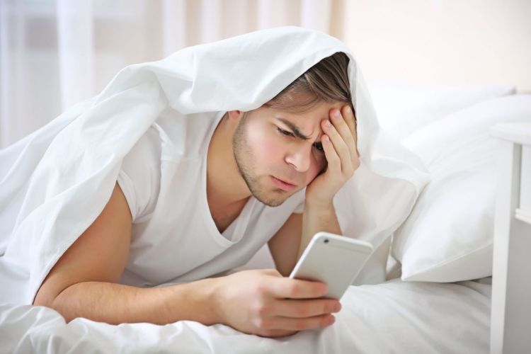 A man with a sheet over head laying on stomach in bed looking at his phone with a disgruntled look on his face